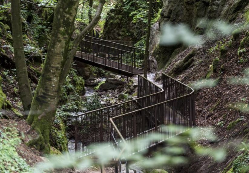 Ein abenteuerlicher Pfad durch die verwunschene Schlucht Ein zickzackförmiger Laufsteg aus Metall führt durch eine bewaldete Schlucht über einen kleinen Bach.