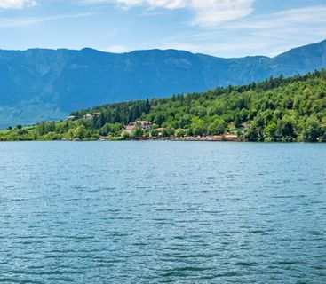 Sommerliche Idylle am glitzernden Bergsee mit einer entspannten Bootsfahrt vor alpiner Kulisse. Ein weites, blaues Gewässer umgeben von dichten grünen Wäldern und einem großen Bergmassiv im Hintergrund, mit einem kleinen Ruderboot auf der Wasserfläche.