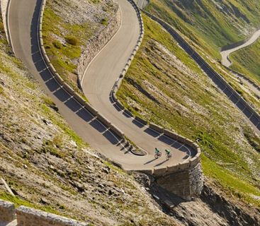 Die ultimative Herausforderung: Sportliche Radtour durch die spektakulären Serpentinen der Alpen Zwei Radfahrer fahren eine steile, kurvenreiche Serpentinenstraße an einem grünen Berghang entlang.