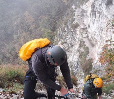 Abenteuer am Fels: Bergsteiger beim Aufstieg in herbstlicher Steilwand. Zwei Personen mit Kletterhelmen und Rucksäcken steigen einen steilen, felsigen Pfad an einem Bergabhang entlang.