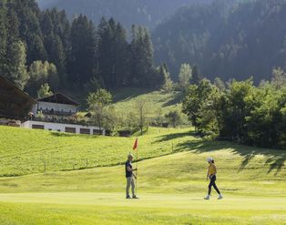 Ein Mann und eine Frau stehen auf einem Golfplatz vor einer alpinen Hügellandschaft mit einem traditionellen Bauernhaus im Hintergrund.
