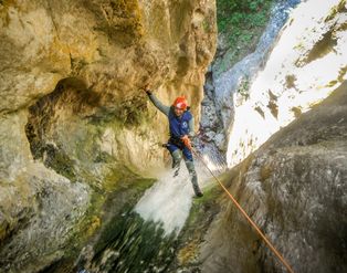 Eine Person in Neoprenanzug und rotem Helm seilt sich bei Canyoning an einer Felswand durch einen Wasserfall ab.