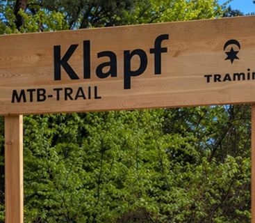 Willkommen am Klapf MTB-Trail – Dein nächstes Mountainbike-Abenteuer in Tramin beginnt hier. Ein hölzernes Hinweisschild vor grünem Wald zeigt die Aufschrift Klapf MTB-Trail in Tramin.