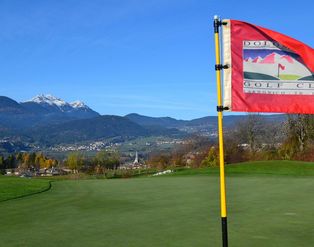 Eine rote Flagge des Dolomiti Golf Clubs weht auf einem Putting Green vor einer Berglandschaft mit einem kleinen Dorf im Tal.