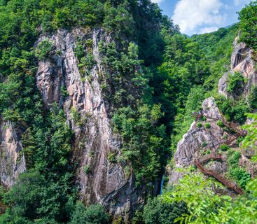 Abenteuerlicher Pfad durch die spektakuläre Felskulisse Eine bewaldete Felsenschlucht mit einer Treppenkonstruktion an einer Klippe, auf der Menschen wandern, sowie einem kleinen Wasserfall im Hintergrund.