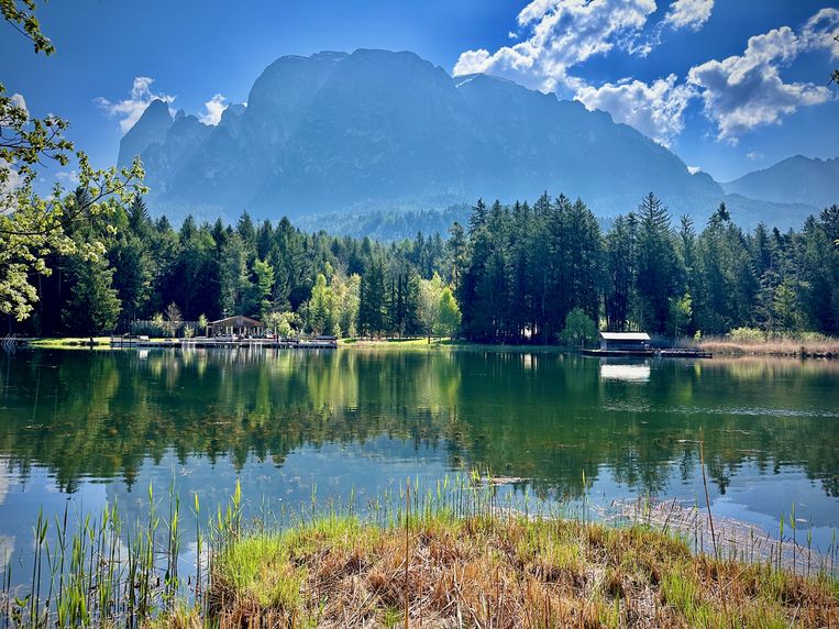 Alpenpanorama am stillen See – majestätische Berge spiegeln sich im klaren Wasser. Bergsee mit Steg und Bootshaus vor einem bewaldeten Bergmassiv unter blauem Himmel.