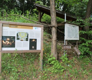 Wanderstation am Rundweg Graun: Ein historischer Lernort im Grünen. Informationstafeln mit Grafiken zur Kalkgewinnung und Wegbeschilderungen stehen auf einer grünen Wiese vor einem Waldstück.