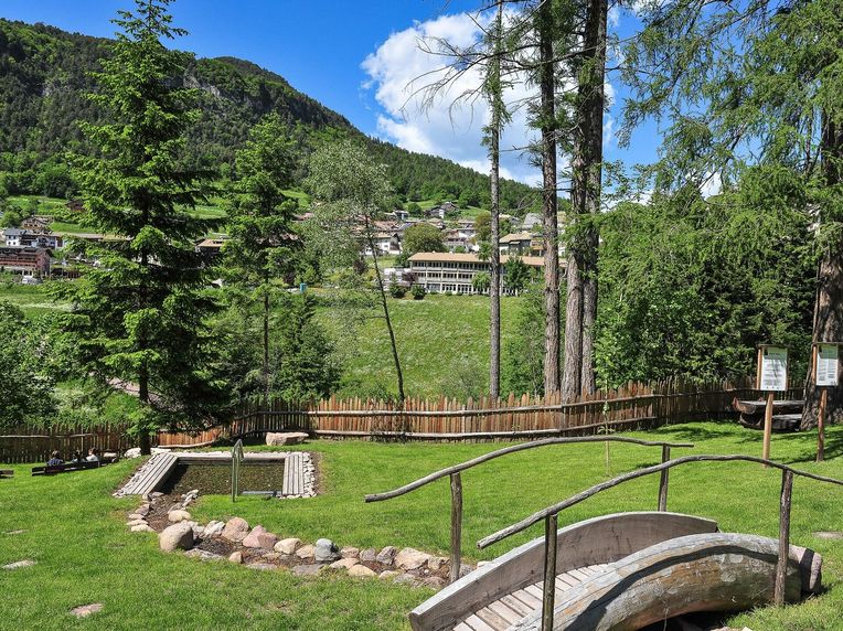 Idyllischer Berggarten mit Bach, Holzbrücke und Blick auf das alpine Dorf Grüne Parkanlage mit Holzbrücke, Bachlauf und kleinem Wasserbecken vor einem Bergdorf und bewaldeten Bergen.