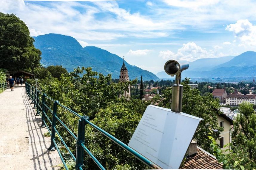 Panoramablick vom Höhenweg auf Kirche, See und majestätische Alpenberge Aussichtspunkt mit Fernrohr über eine Stadt, eine Kirche, den See und die umliegenden Berge; links verläuft ein von Bäumen gesäumter Weg.