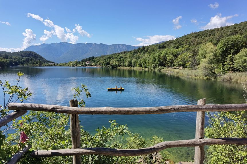 Friedliche Ruderpartie inmitten der alpinen Seenlandschaft an einem sonnigen Sommertag. Blick von einem Holzgeländer auf einen azurblauen Bergsee, auf dem ein kleines gelbes Ruderboot mit zwei Personen im Sonnenschein fährt, im Hintergrund bewaldete Berge.