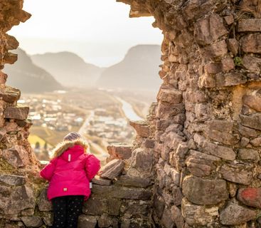 Ein Fenster in die Vergangenheit: Ein kleiner Abenteurer genießt den Ausblick von der alten Burgmauer. Ein Kind in einer leuchtend pinken Winterjacke steht in einer Ruinenöffnung und blickt auf ein Tal hinunter.