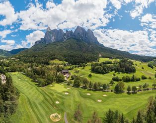Ein Panoramablick auf einen Golfplatz vor dem beeindruckenden, schroffen Bergmassiv des Schlern in Südtirol unter blauem Himmel mit weißen Wolken.