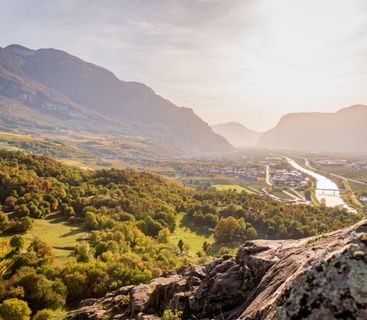 Goldene Herbststimmung im Etschtal: Ein malerischer Ausblick über Weinberge und Flusslauf. Ein Blick von einem felsigen Hügel über ein herbstliches Tal mit einem Fluss, einer kleinen Ortschaft und umgebenden Bergen im warmen Licht der tiefstehenden Sonne.