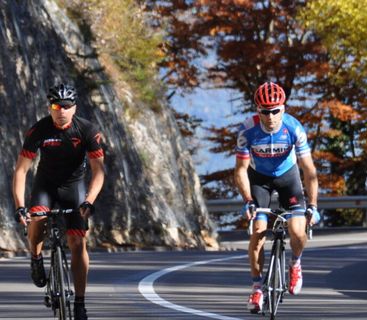 Fahrrad-Abenteuer in herbstlicher Berglandschaft Zwei Radrennfahrer mit Helmen und Sporttrikots fahren auf einer kurvigen Bergstraße entlang eines steilen Felsens und herbstlicher Bäume.