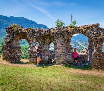 Historische Ausblicke genießen: Abenteuerliche Wanderung durch die antiken Gemäuer mit herrlichem Alpenpanorama. Eine Frau und ein Kind stehen vor einer alten Steinruine mit Torbögen, die einen weiten Blick auf ein Tal mit Feldern, einem Fluss und Bergen im Hintergrund bietet.