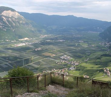 Atemberaubender Fernblick über das Etschtal im Abendlicht Ein weites, von hohen Bergen eingerahmtes Tal, das von oben betrachtet wird. Im Tal erstrecken sich zahlreiche landwirtschaftliche Felder und kleine Ortschaften, im Vordergrund ein metallenes Sicherheitsgeländer.