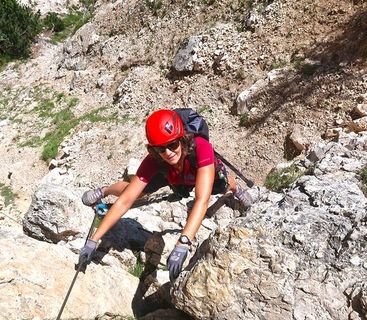 Am Berg zu Hause: Mit Fokus und Kraft am Klettersteig unterwegs. Eine Frau mit rotem Kletterhelm und Handschuhen klettert im Hochgebirge an einem Drahtseil über felsiges Gelände.