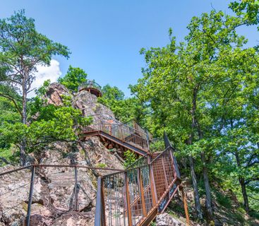 Der Weg zum Gipfelglück: Eine steile Treppe erklimmt den felsigen Aussichtspunkt inmitten grüner Wälder. Eine Metalltreppe führt eine steile Felsformation hinauf, die von grünen Bäumen umgeben ist, bis zu einer kleinen Aussichtsplattform unter einem klaren blauen Himmel.