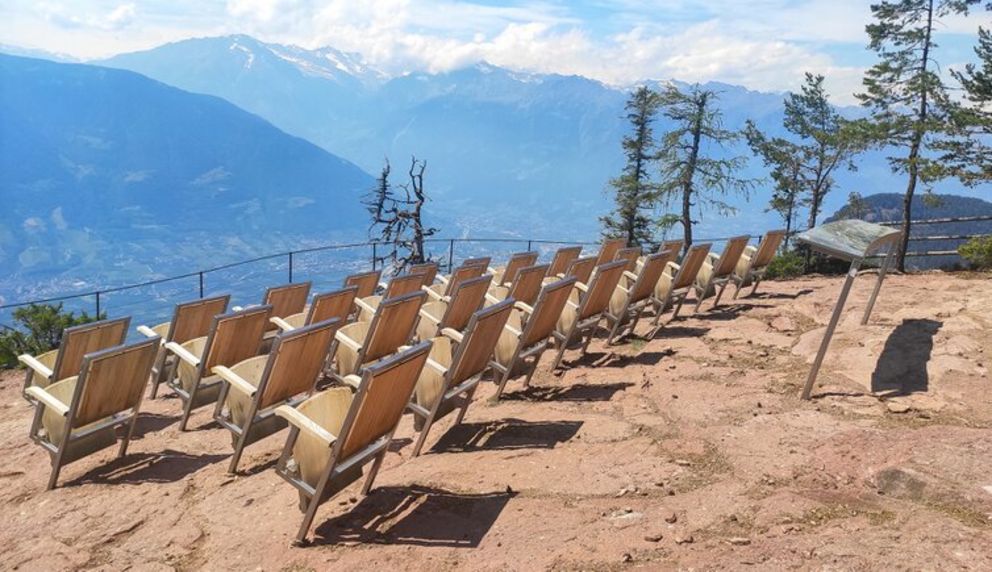 Reihen leerer Holzliegestühle stehen auf einer sonnigen Bergterrasse mit Blick auf ein weites alpines Tal und hohe Berge.