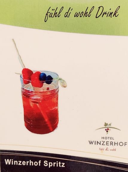 Ein roter Cocktail in einem Glas, garniert mit einer Himbeere und einer Blaubeere am Spieß, vor einem Werbehintergrund für den Winzerhof Spritz.