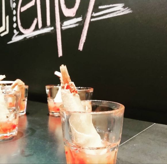 Glas mit rotem Getränk und Garnelen am Rand vor einer Tafel mit Kreideschrift