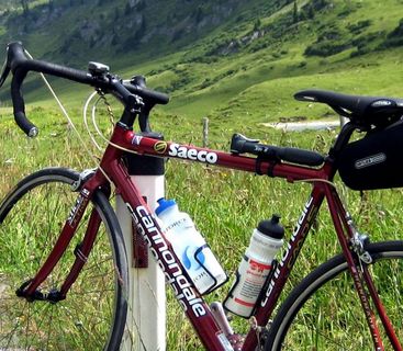 Bereit für den Anstieg – Das Reise-Rennrad in alpiner Kulisse. Ein rotes Rennrad mit Satteltasche und Trinkflaschen steht an einer Berghangstraße neben einer Leitpfosten.