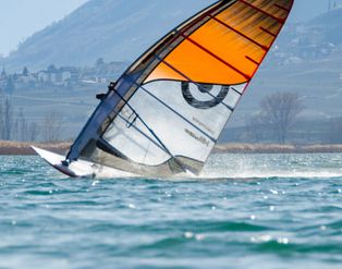 Ein Windsurfer mit einem orangefarbenen Segel fährt bei sonnigem Wetter auf einem See vor einer bergigen Kulisse.