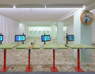 Ein Innenraum eines Schießstandes mit mehreren grünen Schießtischen, digitalen Monitoren zur Zielanzeige und Scheiben im Hintergrund.