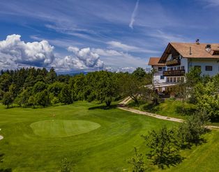 Ein sonniger Golfplatz mit einem grünen Fairway, einem Bunker und einem Gebäude im alpenländischen Stil im Hintergrund vor einem blauen Himmel mit Wolken.
