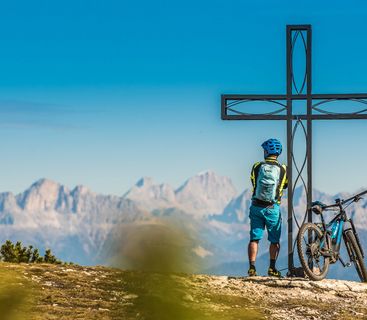 Der verdiente Ausblick: Gipfelglück mit dem E-Bike in den Alpen. Ein Mountainbiker steht mit seinem E-Bike neben einem Gipfelkreuz vor einem weitläufigen Gebirgspanorama bei sonnigem Wetter.